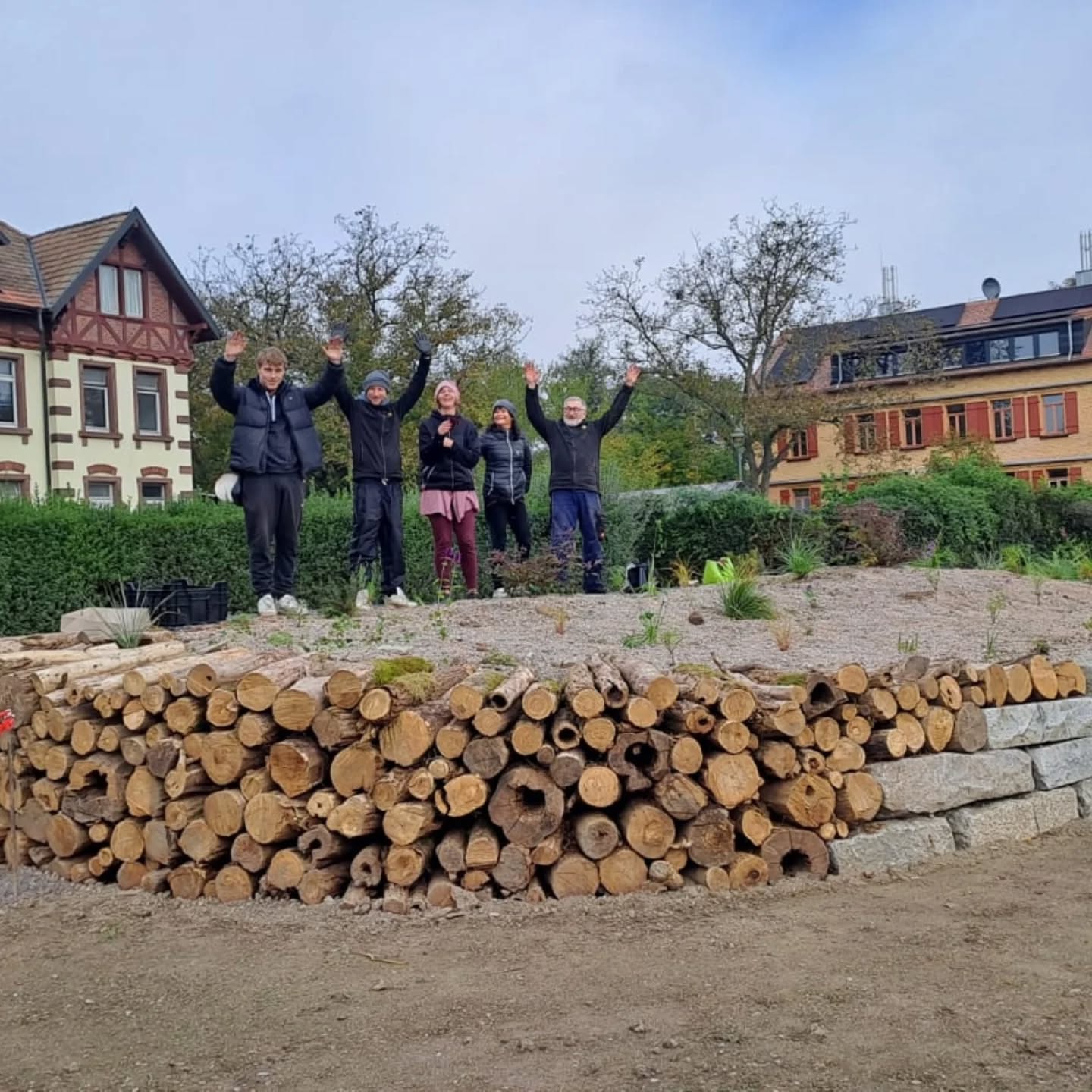 Dieser Alternativtext wurde KI-generiert: Mehrere Personen stehen im Freien vor einem Gebäude und heben die Arme in die Luft. Im Vordergrund befindet sich ein Holzstapel und eine bepflanzte Anhöhe.