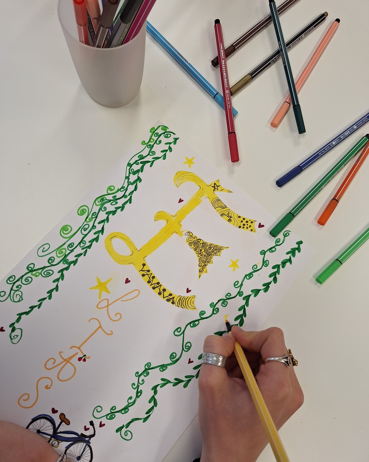 Dieser Alternativtext wurde KI-generiert: Ein farbenfrohes Bild zeigt eine Hand, die mit einem Stift auf ein Blatt Papier malt. Auf dem Papier befindet sich eine detaillierte, verzierte Zeichnung mit floralen Elementen und abstrakten Formen.