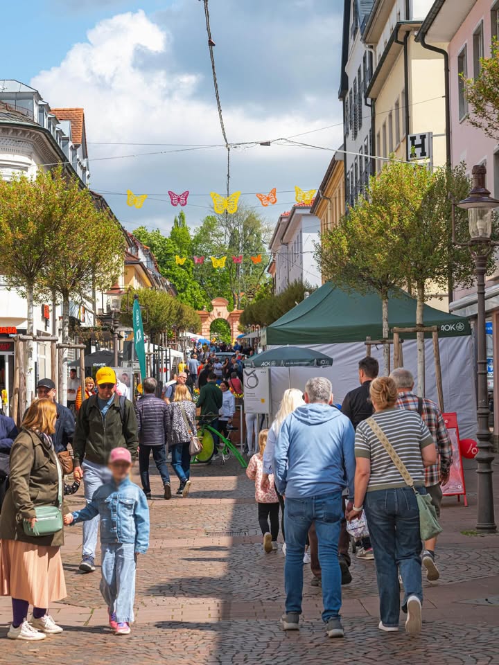 Dieser Alternativtext wurde KI-generiert: Eine belebte Straße mit Geschäften und Bäumen. Menschen gehen auf dem Pflasterweg durch die Stadt, einige sind mit Kindern unterwegs.
