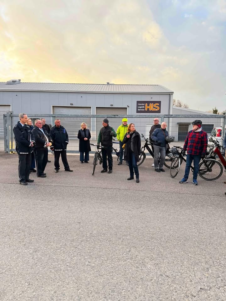Dieser Alternativtext wurde KI-generiert: Eine Gruppe von Personen steht und sitzt vor einem Gebäude und mehreren Fahrrädern. Im Hintergrund ist ein grauer Himmel zu sehen.