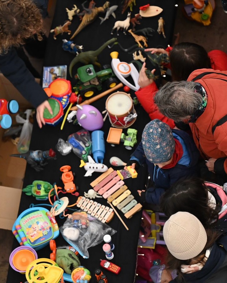 Dieser Alternativtext wurde KI-generiert: Eine Ansammlung von Spielzeug liegt auf einer dunklen Oberfläche. Mehrere Hände sind im Bild zu sehen, die sich in der Nähe des Spielzeugs befinden.