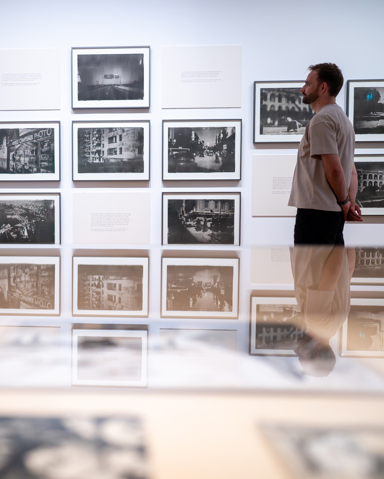 Ein Mensch in einem hellen T-Shirt und dunklen Shorts steht mit verschränkten Händen vor einer Wand, die mit einem Raster aus schwarz-weißen Fotografien und Texttafeln bedeckt ist. Im unteren Bildbereich ist die Spiegelung der Person und der Ausstellung auf einer glänzenden Fläche zu sehen. (KI-Alternativtext)