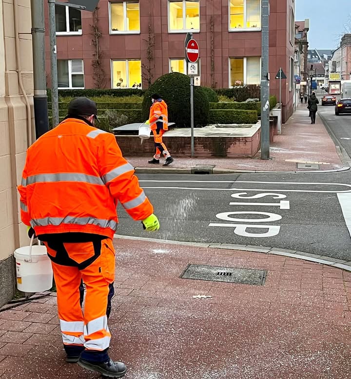 Dieser Alternativtext wurde KI-generiert: Eine Straßenansicht zeigt Personen bei der Arbeit an einer Kreuzung. Auf dem Boden ist ein großes weißes 'STOP'-Schild zu sehen.