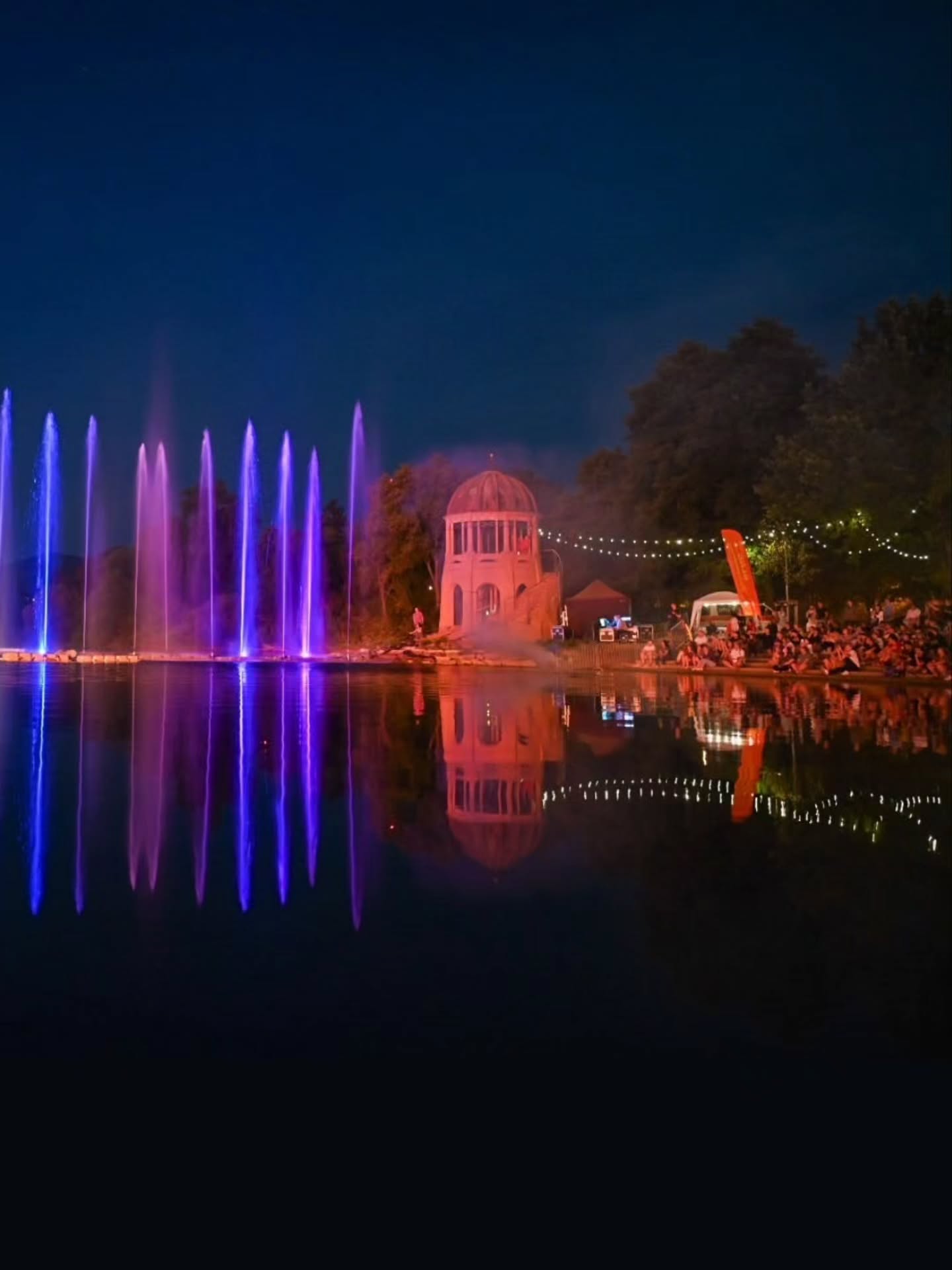 Bei Nacht findet am Ufer eines Gewässers eine Licht- und Wassershow statt. Links sprudeln mehrere Fontänen, die in Blau und Lila beleuchtet sind, während in der Mitte ein kuppelförmiges Gebäude in rotem Licht erstrahlt. Rechts sitzt eine große Gruppe von Menschen am Wasser und beobachtet die Show, wobei sich die Lichter und Strukturen im ruhigen Wasser spiegeln. (KI-Alternativtext)