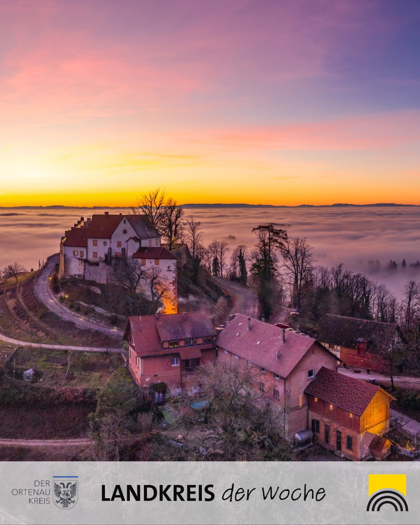 Dieser Alternativtext wurde KI-generiert: Ein Gebäudeensemble thront auf einem Hügel in nebliger Landschaft während eines Sonnenaufgangs. Das Bild zeigt warme Farben und eine friedliche Atmosphäre.