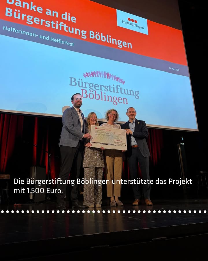 Dieser Alternativtext wurde KI-generiert: Eine Gruppe von Menschen steht auf einer Bühne vor einem großen Bildschirm. Sie halten einen symbolischen Scheck und lächeln in die Kamera.