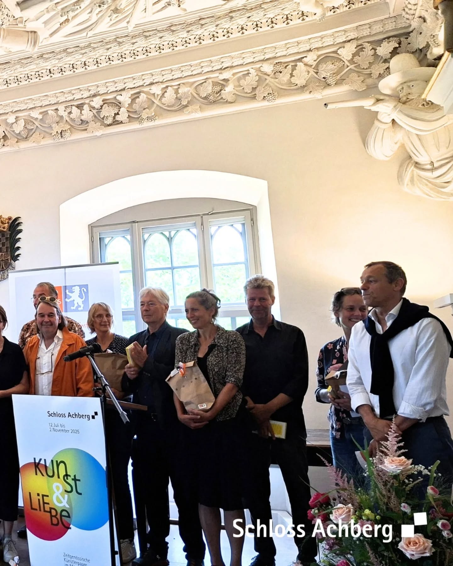 Eine Gruppe von Leuten steht vor einem Poster mit der Aufschrift Schloss Achberg. Der Raum hat eine dekorative Decke.