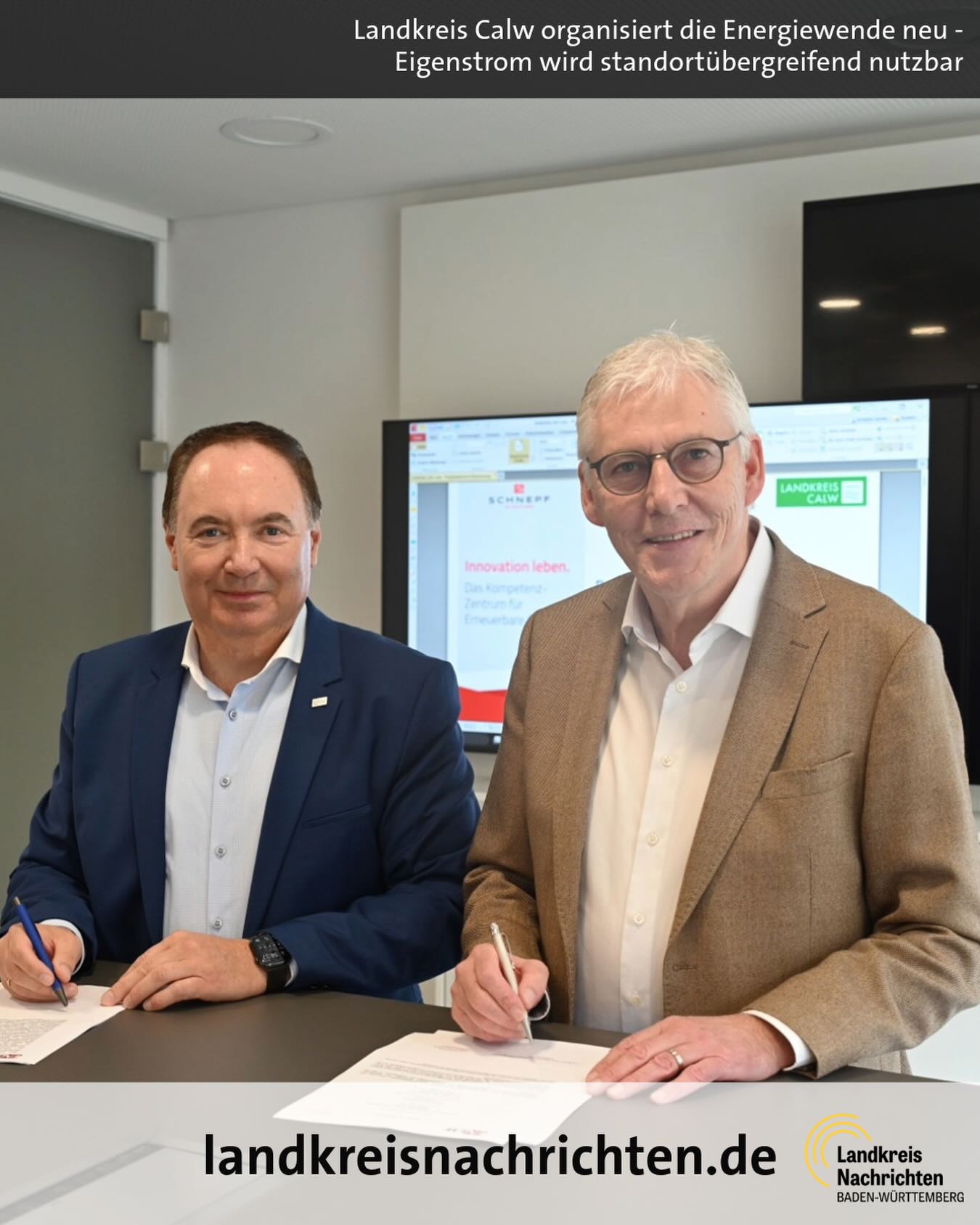 Dieser Alternativtext wurde KI-generiert: Zwei Personen in formeller Kleidung sitzen an einem Tisch und lächeln in die Kamera. Im Hintergrund ist ein Bildschirm mit Text und Logo zu sehen.