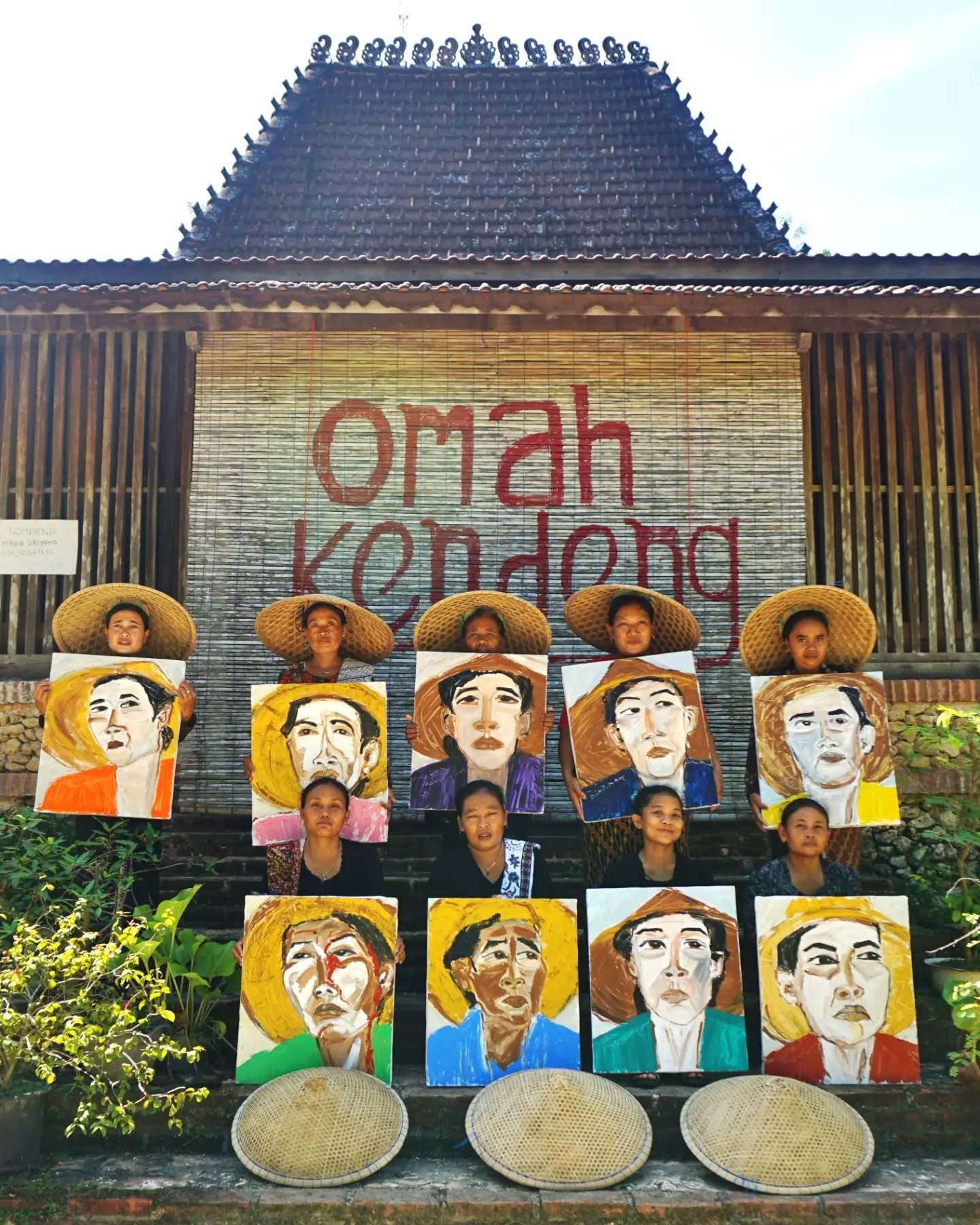 Mehrere Personen mit Strohhüten halten Gemälde von Gesichtern vor sich, dahinter ein Gebäude mit der Aufschrift „Omah Kendeng“. Drei weitere Strohhüte liegen vorne auf dem Boden. (KI-Alternativtext)