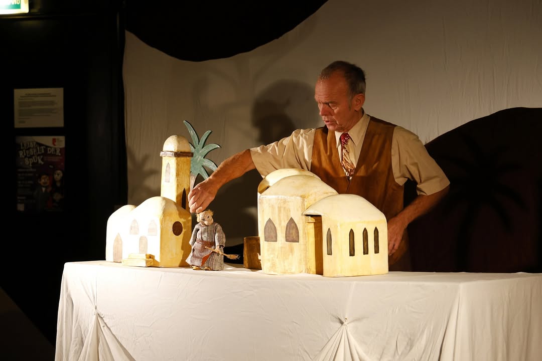 Dieser Alternativtext wurde KI-generiert: Eine Person in einem Hemd und einer Weste präsentiert eine Krippenszene mit Figuren vor einem dunklen Hintergrund. Die Szene steht auf einem mit Stoff bedeckten Tisch.