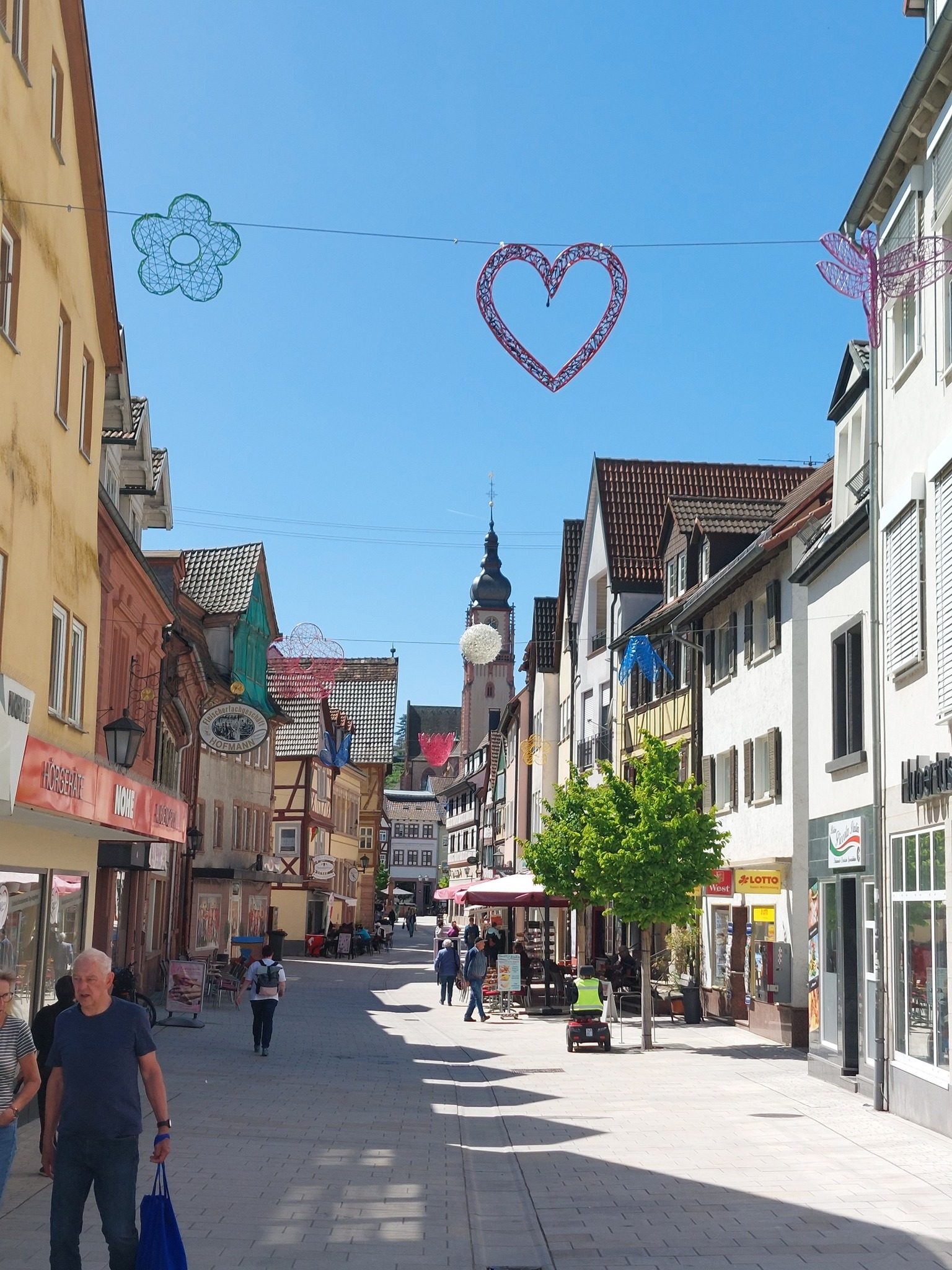 Dieser Alternativtext wurde KI-generiert: Eine sonnige Straßenszene mit bunten Dekorationen, die zwischen den Gebäuden hängen. Die Straße ist von historischen Häusern und einer Kirche gesäumt.