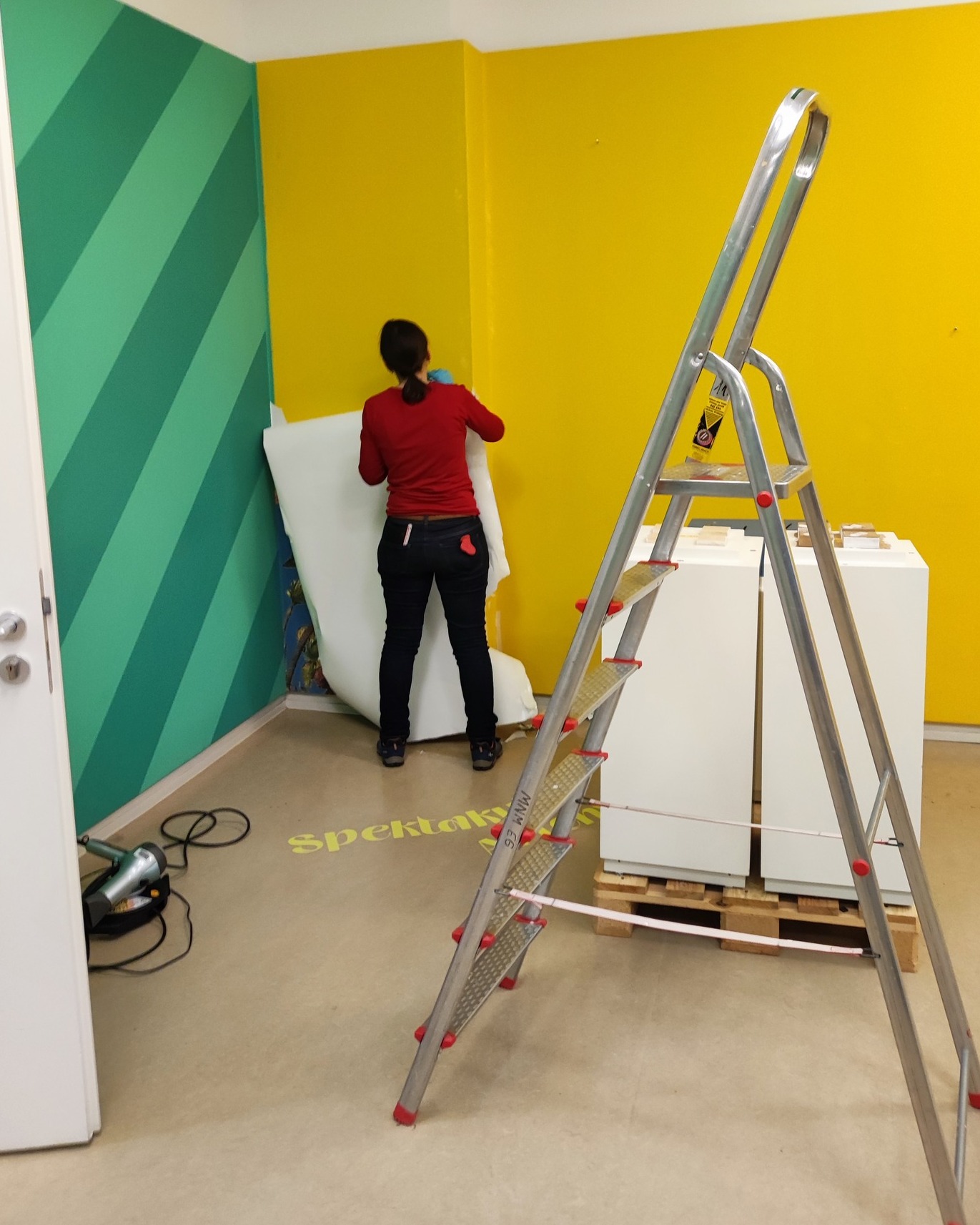 Dieser Alternativtext wurde KI-generiert: Ein Innenraum mit farbigen Wänden, an denen gearbeitet wird. Eine Person steht auf einer Leiter und hängt etwas auf.