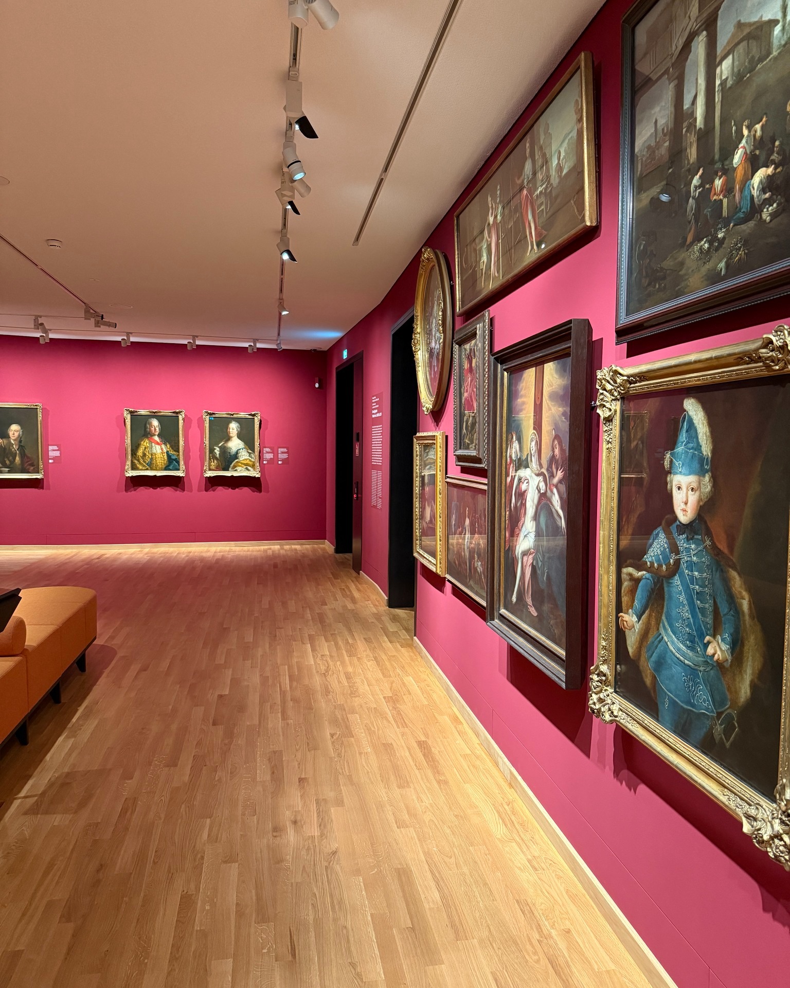 Ausstellungssaal mit leuchtend pinken Wänden, an denen mehrere gerahmte Gemälde aus vergangenen Epochen hängen, darunter Porträts und Szenen mit Figuren; Holzboden, Deckenstrahler und eine orangefarbene Sitzbank im Vordergrund. (KI-Alternativtext)