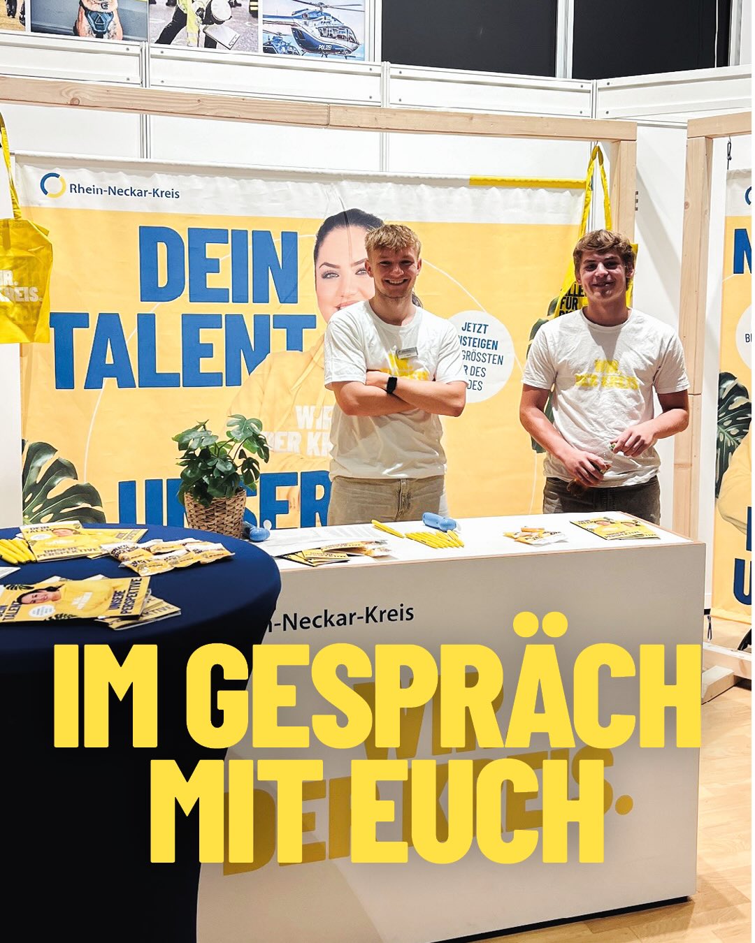 Zwei Personen stehen lächelnd hinter einem weißen Stand mit der großen Aufschrift „IM GESPRÄCH MIT EUCH.“. Im Hintergrund hängt ein gelbes Banner mit dem Text „DEIN TALENT“ und dem Logo des Rhein-Neckar-Kreises. Auf dem Stand und einem runden Tisch daneben liegen Flyer und Stifte bereit. (KI-Alternativtext)