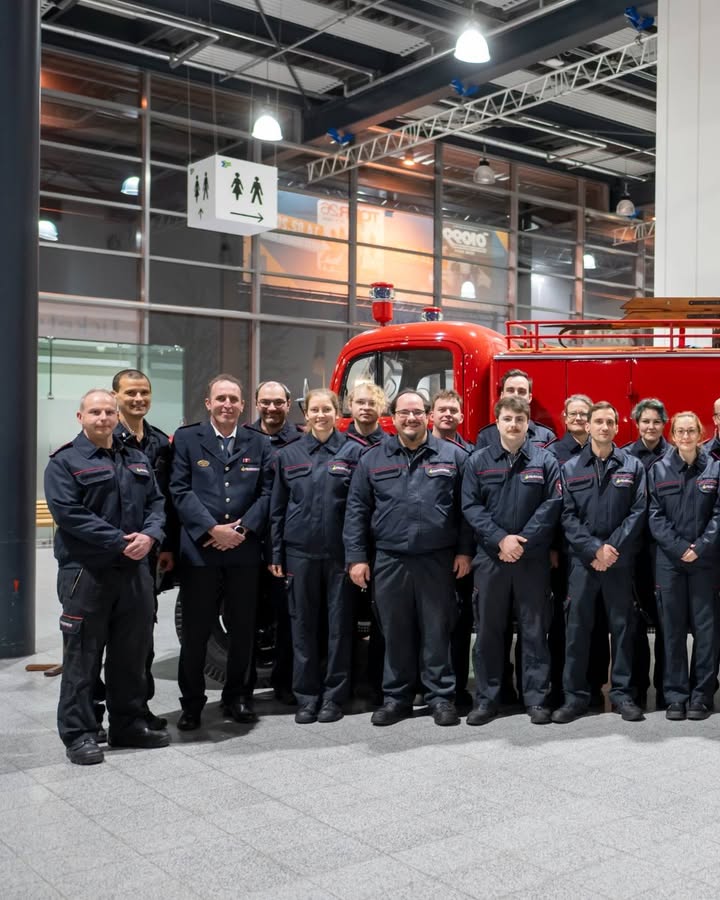 Dieser Alternativtext wurde KI-generiert: Eine Gruppe von Personen in dunkler Arbeitskleidung steht vor einem roten Feuerwehrauto in einer modernen Halle. Im Hintergrund sind weitere Personen und Schilder zu sehen.
