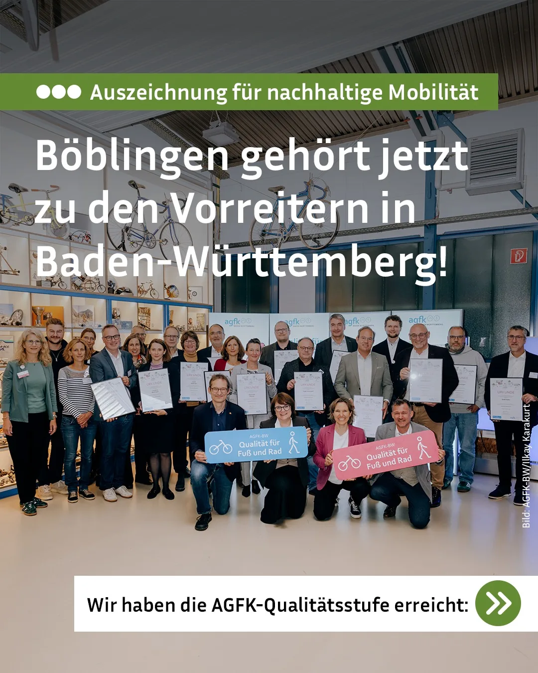 Eine Gruppe von Menschen steht und kniet in einem Raum mit Fahrrädern an der Wand. Sie halten Urkunden sowie blaue und rosa Schilder mit dem Text „AGFK-BW Qualität für Fuß und Rad“. Der eingeblendete Text lautet: „Auszeichnung für nachhaltige Mobilität – Böblingen gehört jetzt zu den Vorreitern in Baden-Württemberg! Wir haben die AGFK-Qualitätsstufe erreicht.“ (KI-Alternativtext)