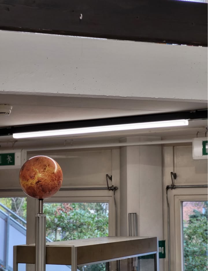 Dieser Alternativtext wurde KI-generiert: Ein runder, bunt gefärbter Globus steht auf einem Fuß vor einer Fensterfront. Darüber befindet sich eine lange, helle Leuchte unter der Decke.