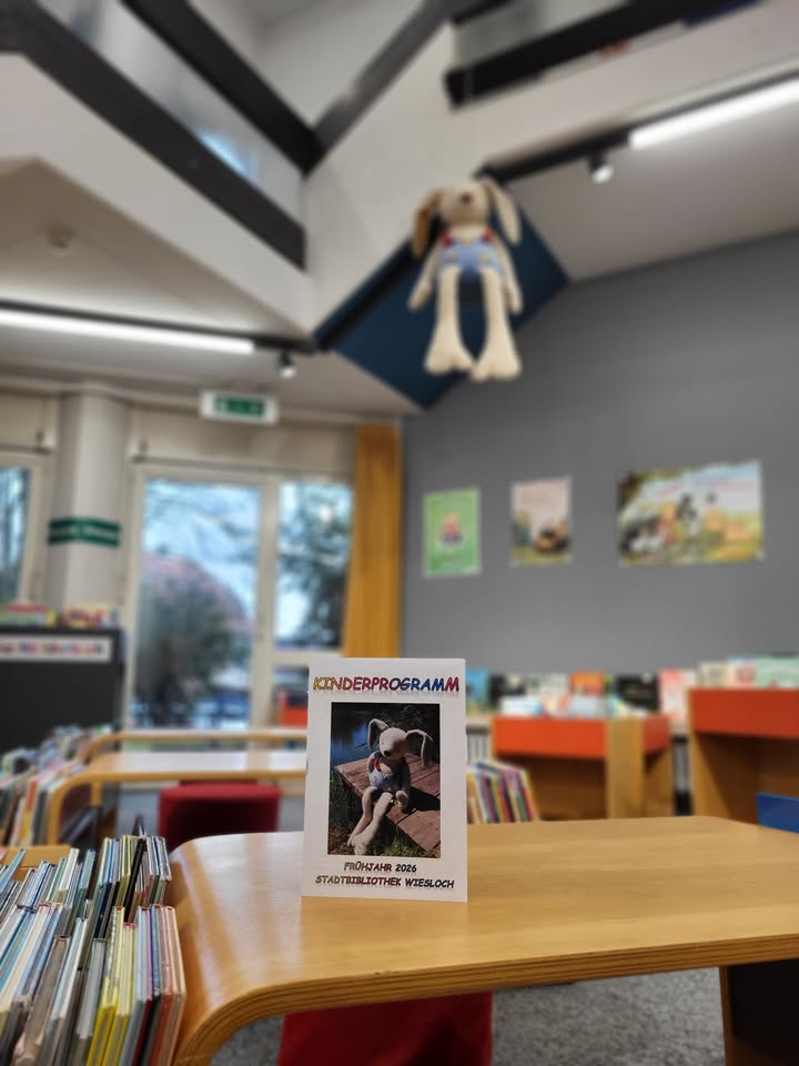 Dieser Alternativtext wurde KI-generiert: Ein Tisch mit Broschüren und Büchern steht im Vordergrund. Im Hintergrund ist ein Raum mit Fenstern und Dekorationen zu sehen.