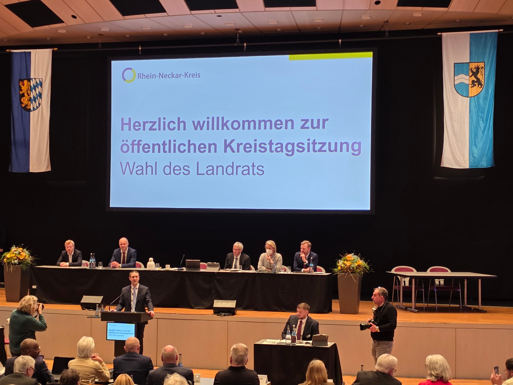 Dieser Alternativtext wurde KI-generiert: Eine Präsentation mit dem Titel 'Herzlich willkommen zur öffentlichen Kreistagssitzung Wahl des Landrats' wird auf einer großen Leinwand gezeigt. Menschen sitzen an einem Tisch auf einer Bühne, während andere im Publikum anwesend sind.