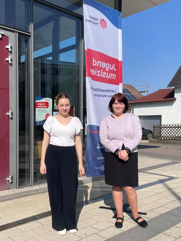 Dieser Alternativtext wurde KI-generiert: Zwei Personen stehen vor einem Gebäude mit Glasfassade und einem Banner. Im Hintergrund ist ein Garten mit Bäumen und Büschen zu sehen.