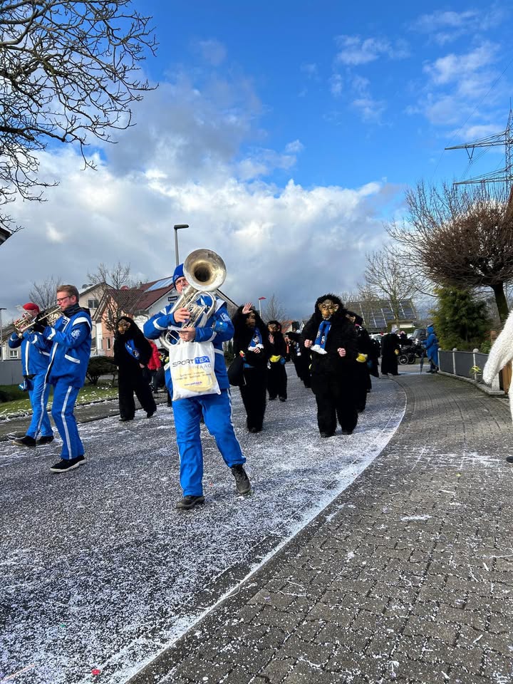 Dieser Alternativtext wurde KI-generiert: Eine Gruppe von Personen marschiert auf einem Weg, einige spielen Blechblasinstrumente. Im Hintergrund sind Häuser und ein bewölkter Himmel zu sehen.