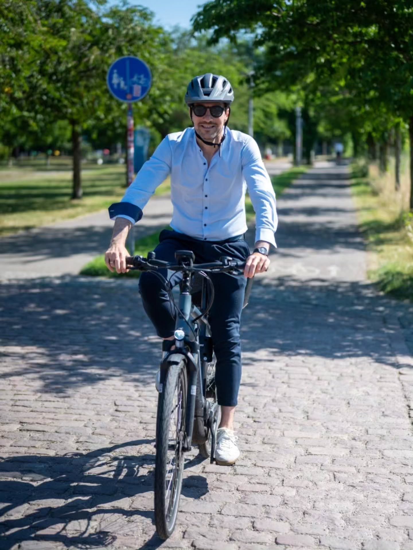 Eine Person mit Helm und Sonnenbrille fährt lächelnd ein Fahrrad auf einem gepflasterten Weg zwischen Bäumen. Im Hintergrund ist ein blaues Verkehrsschild mit Rad- und Gehwegsymbolen zu sehen. (KI-Alternativtext)