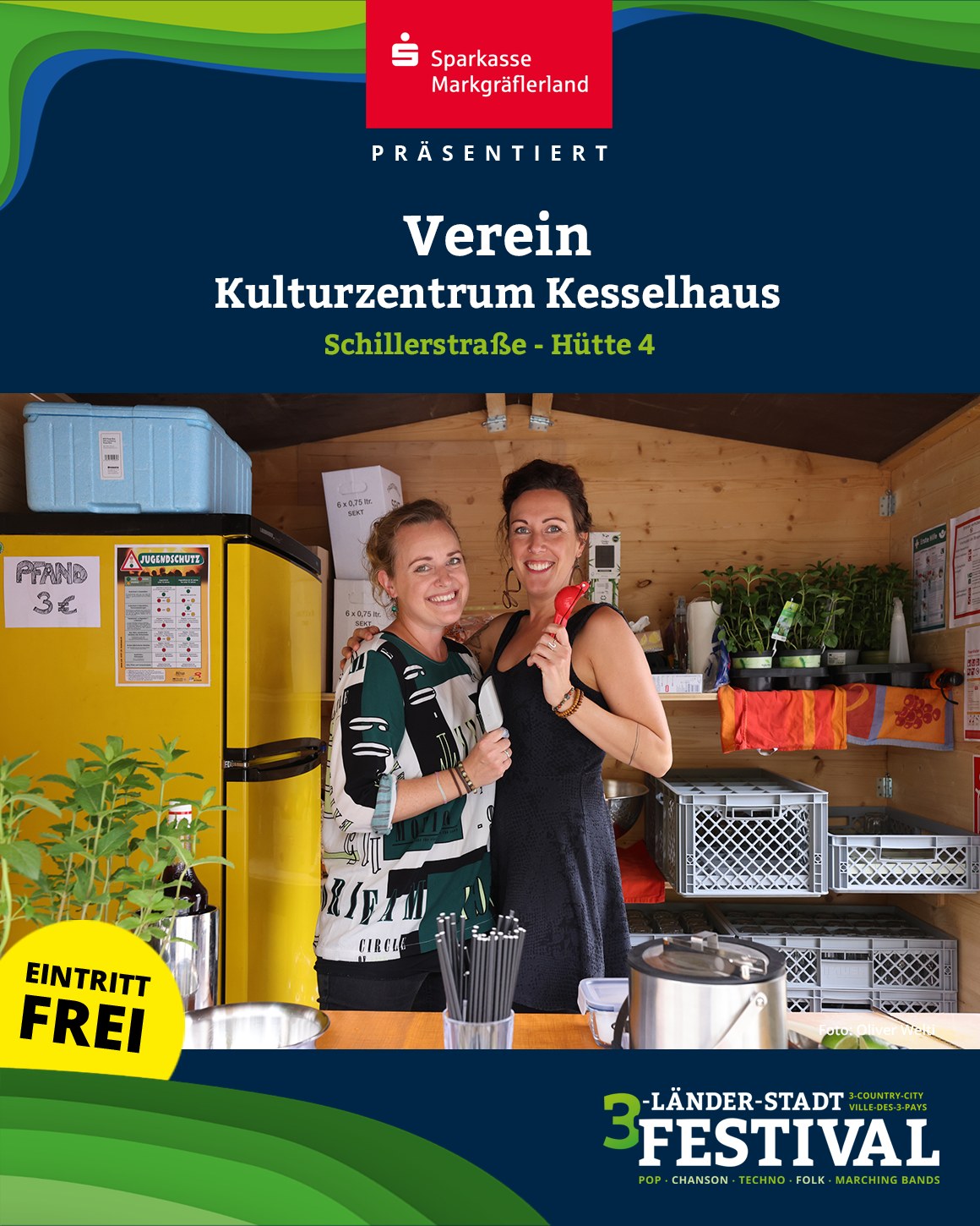 Zwei Personen stehen lächelnd vor einem Stand mit verschiedenen Gegenständen und Pflanzen vor der Werbung für ein Festival.