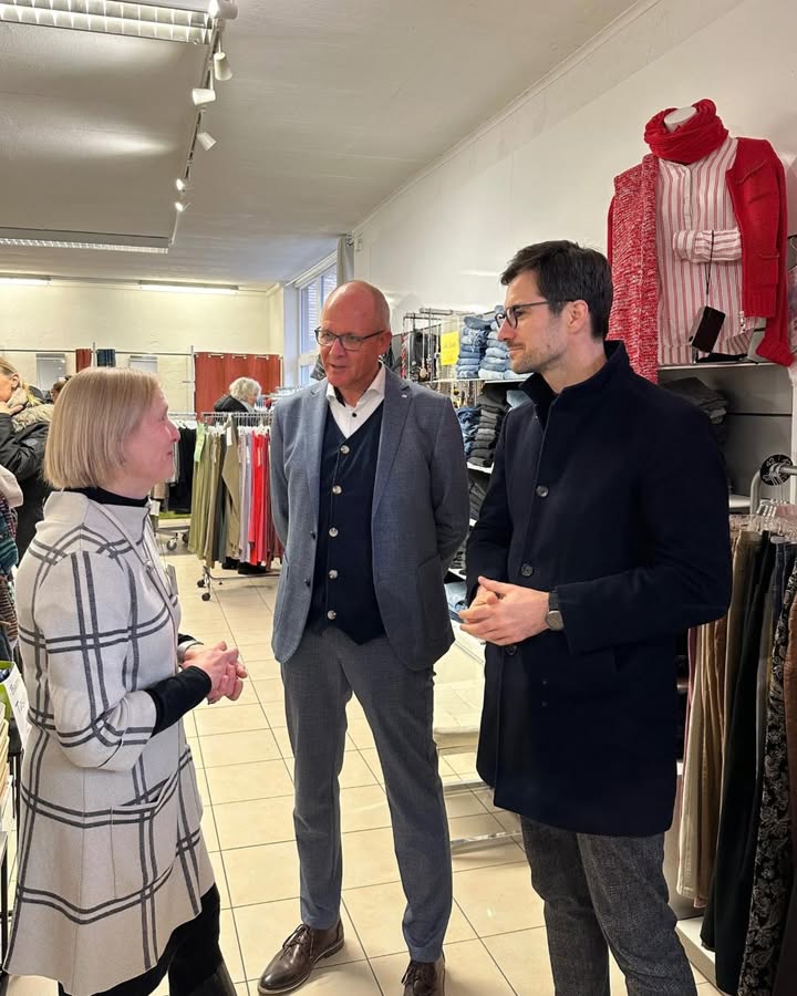 Dieser Alternativtext wurde KI-generiert: Mehrere Personen stehen in einem Geschäft und unterhalten sich. Im Hintergrund sind Kleidung und Regale zu sehen.