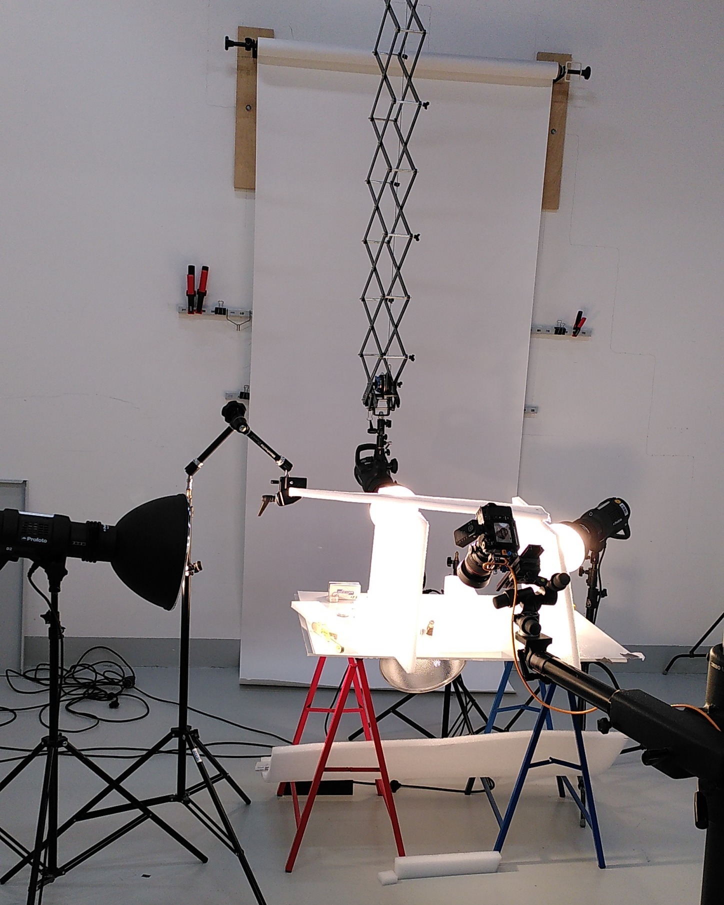 Dieser Alternativtext wurde KI-generiert: Ein Fotostudio mit mehreren Lampen, Stativen und einer Beleuchtungseinrichtung.  Im Zentrum steht ein Gegenstand auf einem Tisch, der beleuchtet wird.