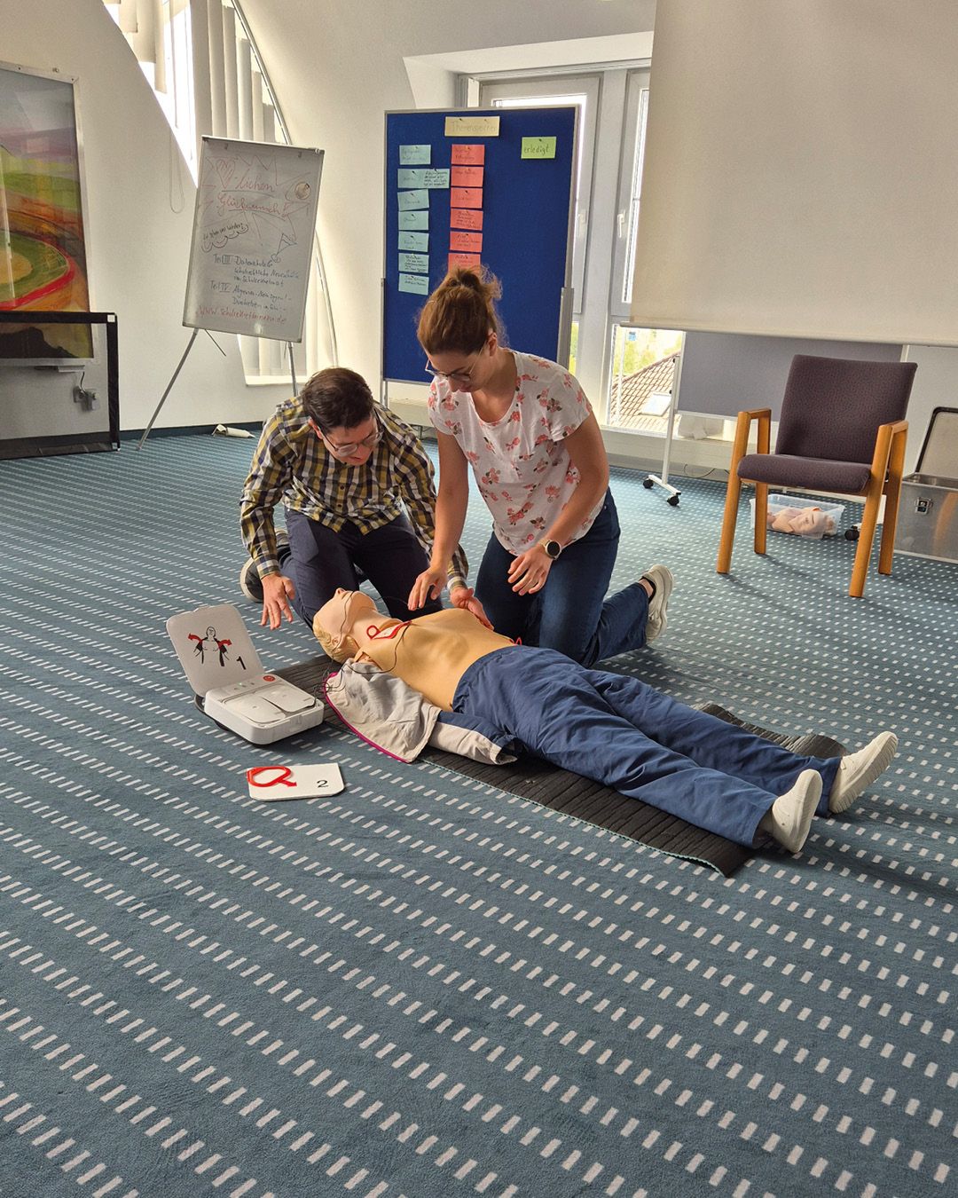 Zwei Personen knien auf dem Boden neben einer liegenden Reanimationspuppe und üben Herzdruckmassage. Ein geöffneter Defibrillator liegt neben dem Kopf der Puppe. Im Hintergrund stehen ein Whiteboard und eine Pinnwand. (KI-Alternativtext)