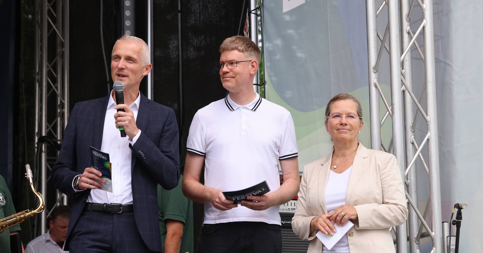 Drei Menschen stehen auf einer Bühne. Links im Bild hält jemand ein Mikrofon und eine Broschüre in der Hand. In der Mitte hält ein Mann eine Broschüre in der Hand und lächelt. Rechts im Bild lächelt eine Frau, die eine Broschüre in der Hand hält.
