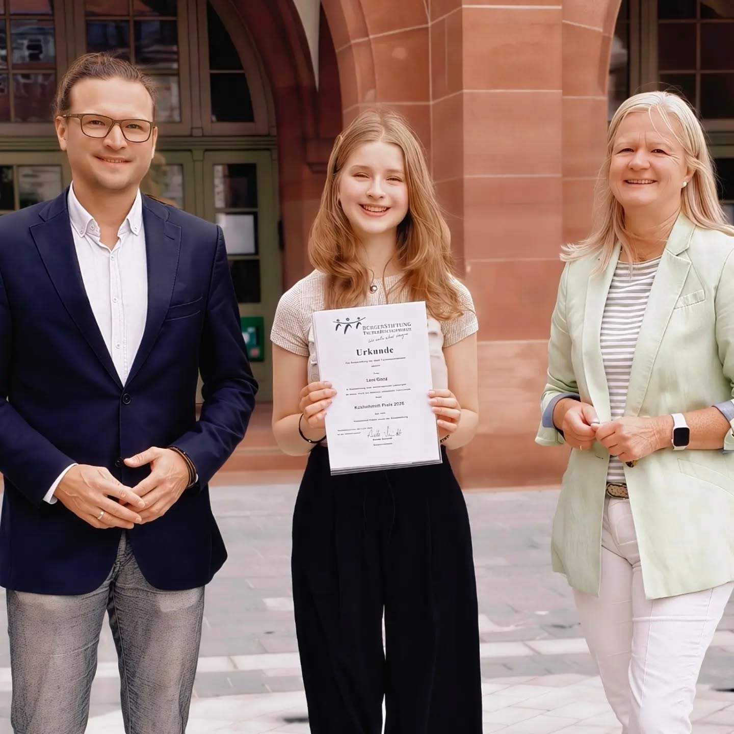 Drei Personen stehen nebeneinander vor einem Gebäude. Die Person in der Mitte hält eine Urkunde der Bürgerstiftung Tuttingen für den Kulturschirm Preis 2026 hoch. Links und rechts stehen jeweils eine weitere Person in Jacke. (KI-Alternativtext)