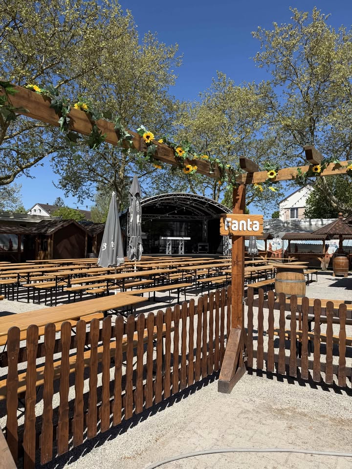 Dieser Alternativtext wurde KI-generiert: Ein hölzerner Rankbogen, geschmückt mit Sonnenblumen und Grünpflanzen, steht vor einer Bühne im Freien. Im Vordergrund befindet sich ein Holzzaun und eine Fläche mit Tischen und Stühlen.