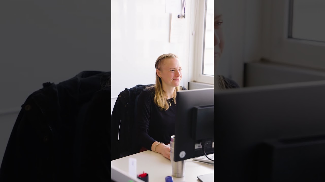 Dieser Alternativtext wurde KI-generiert: Eine Person sitzt an einem Schreibtisch vor einem Computer. Es ist ein Innenraum mit einem Fenster im Hintergrund zu sehen.