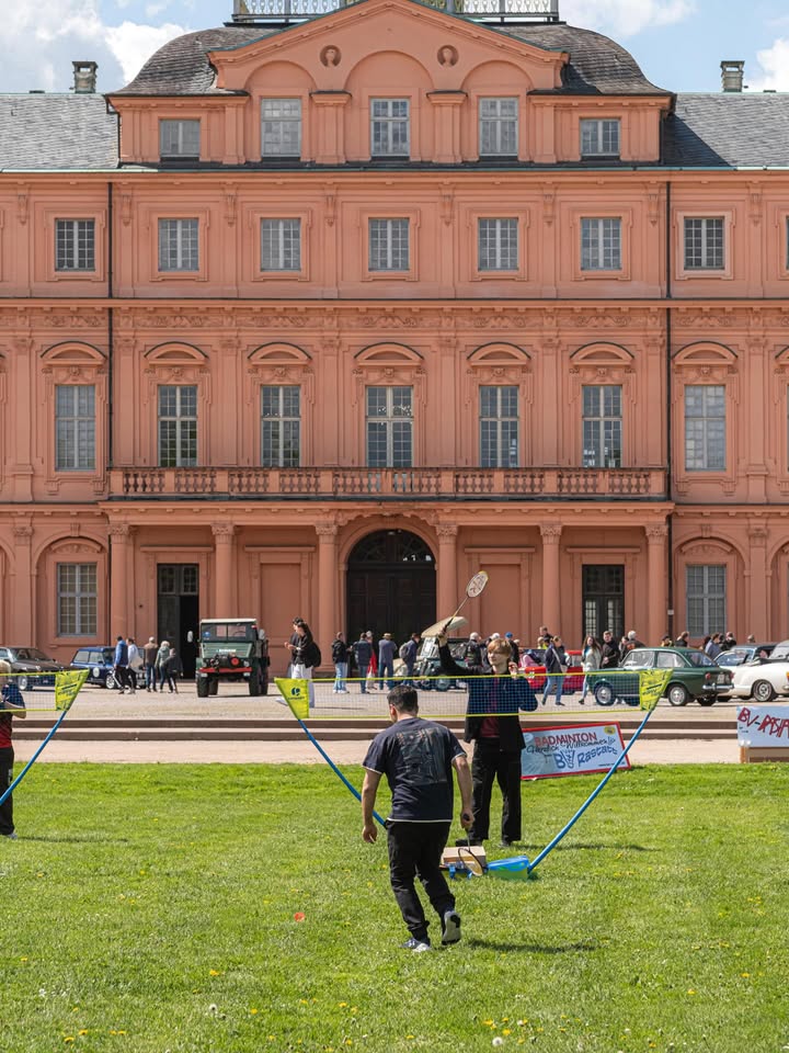 Dieser Alternativtext wurde KI-generiert: Ein großes, helles Gebäude mit vielen Fenstern. Vor dem Gebäude sind Menschen und Fahrzeuge auf einer Rasenfläche zu sehen.