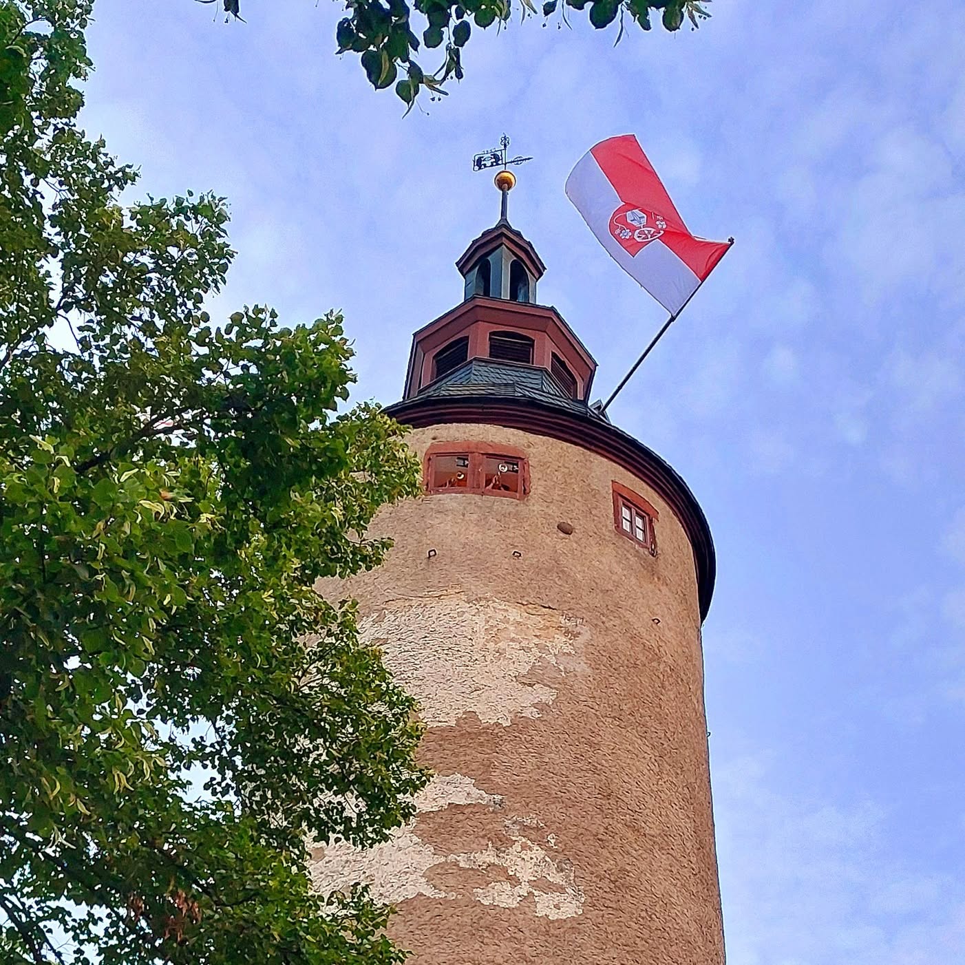 Turm mit Fahne vor blauem Himmel. Ein Teil des Turms ist von Bäumen verdeckt.