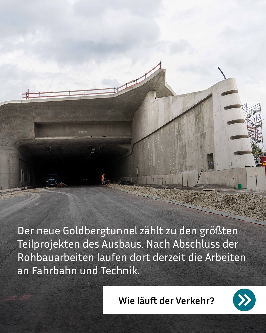 Ein neuer Tunnelbau aus Beton mit einer asphaltierten Fahrbahn und einem dunklen LKW im Inneren. Eine Person in orangefarbener Warnkleidung steht am Rand der Fahrbahn neben Baugerüst und Sandhaufen. Der eingeblendete Text lautet: „Der neue Goldbergtunnel zählt zu den größten Teilprojekten des Ausbaus. Nach Abschluss der Rohbauarbeiten laufen dort derzeit die Arbeiten an Fahrbahn und Technik.“ Darunter befindet sich ein Button mit dem Text „Wie läuft der Verkehr?“ und einem Pfeil-Symbol. (KI-Alternativtext)