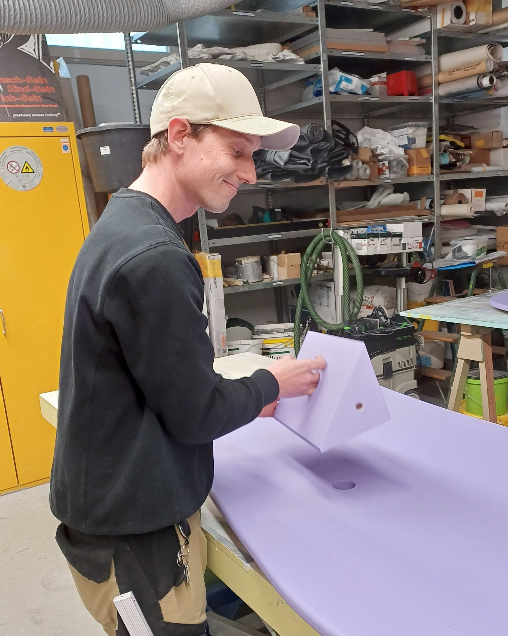 Dieser Alternativtext wurde KI-generiert: Eine Person arbeitet in einer Werkstatt an einem lila Objekt. Im Hintergrund sind Regale mit Werkzeugen und Materialien zu sehen.