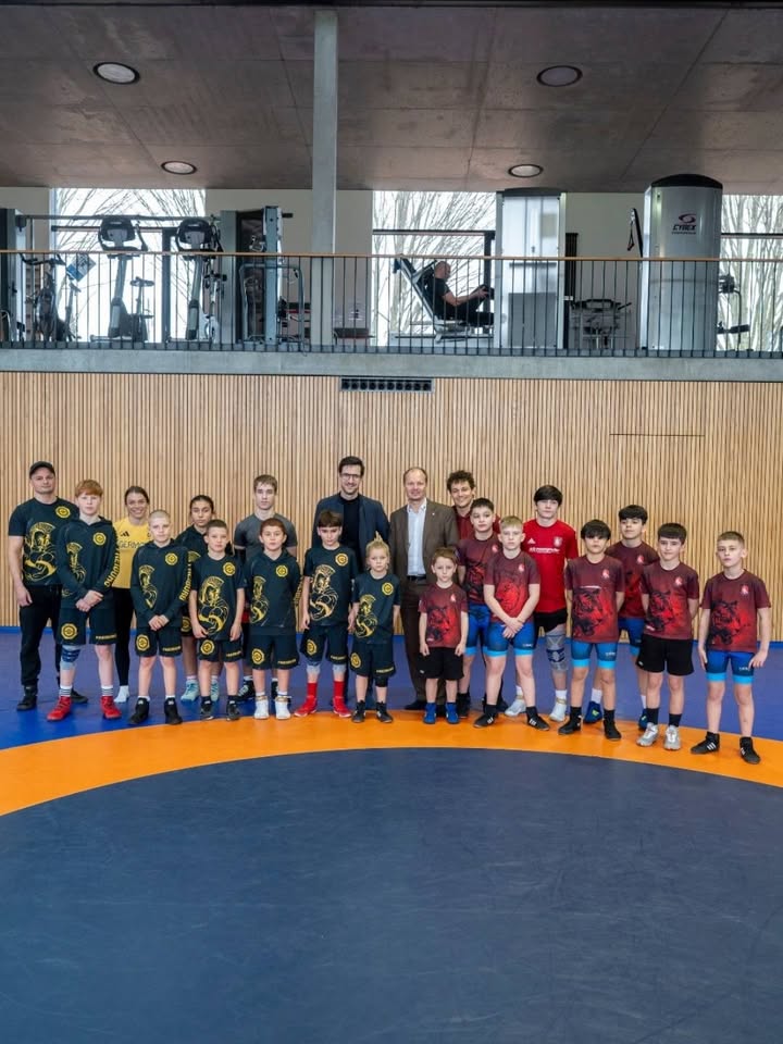 Dieser Alternativtext wurde KI-generiert: Eine Gruppe von Personen steht in einer Halle mit Sportgeräten und großen Fenstern vor einer Holzwand. Im Vordergrund posiert eine Mannschaft auf einer blauen Matte.