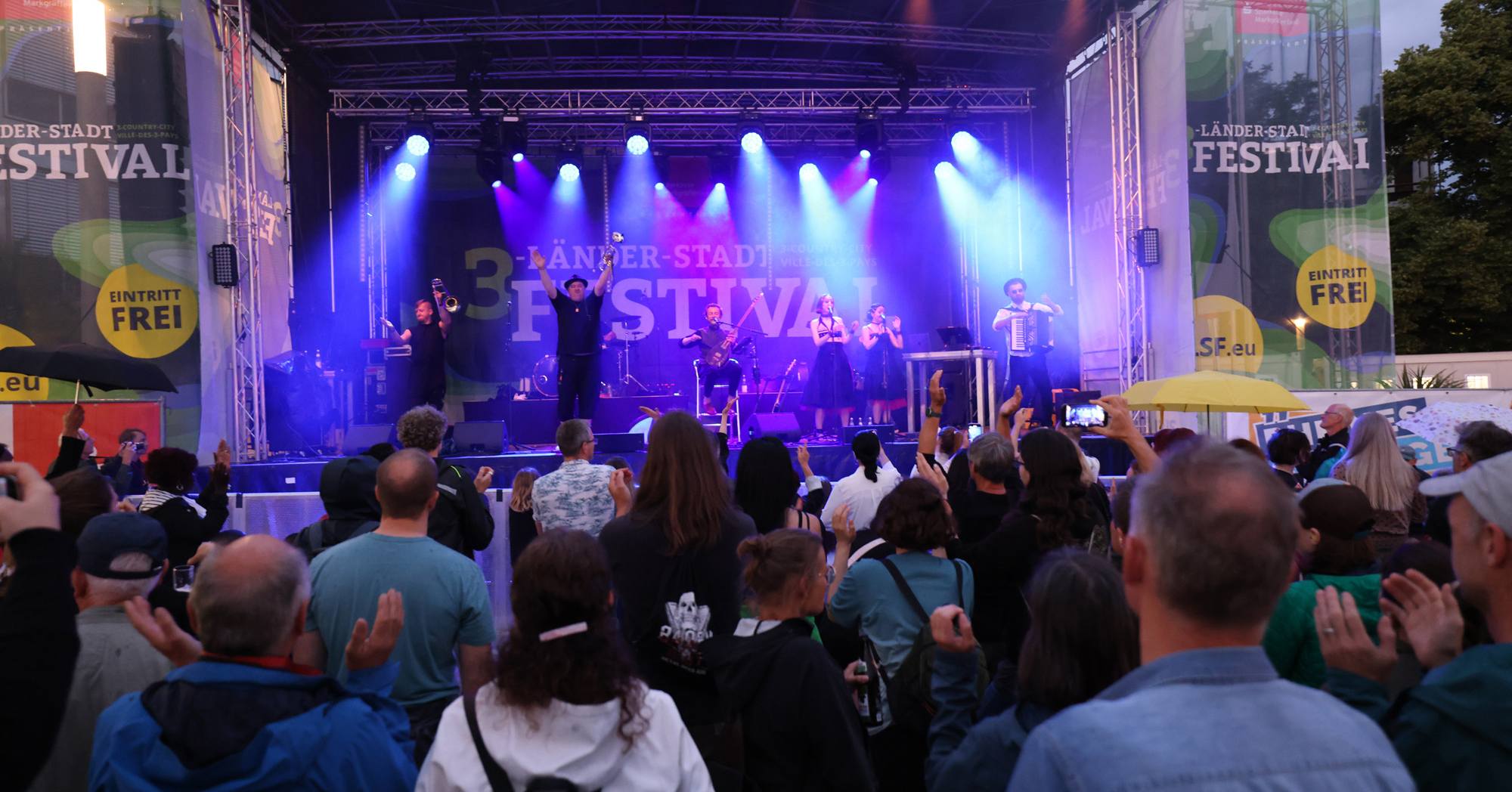 Auf einer Bühne mit Lichtern stehen Menschen und spielen Musik. Zuschauer*innen klatschen und feiern die Musik.
