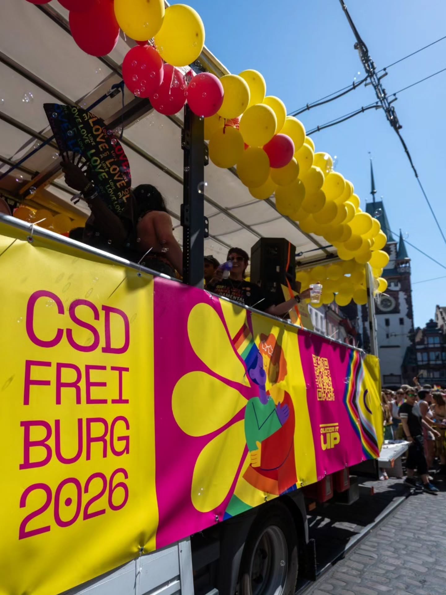 Ein Parade-Wagen mit gelb-roten Ballons und einem Banner, das „CSD FREIBURG 2026“ zeigt, sowie eine Illustration von zwei sich umarmenden Figuren und einem QR-Code. Personen auf dem Wagen halten Fächer mit der Aufschrift „LOVE“ und trinken Getränke, im Hintergrund sind Zuschauer und ein historisches Gebäude zu sehen. (KI-Alternativtext)