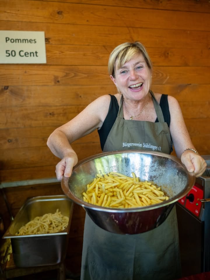 Eine Person mit kurzen blonden Haaren und Schürze hält eine große Metallschüssel mit Pommes Frites. Im Hintergrund ein Schild mit „Pommes 50 Cent“ an einer Holzwand. (KI-Alternativtext)