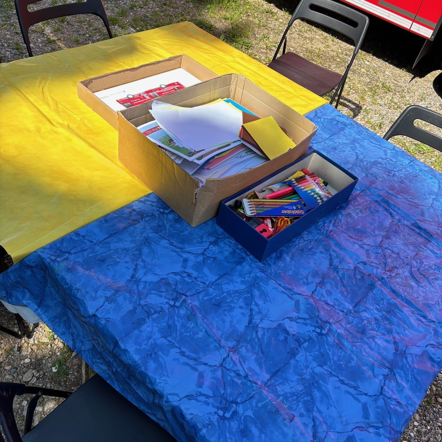 Ein Tisch mit gelber und blauer Tischdecke. Auf dem Tisch stehen eine Kiste mit Papier und eine Schachtel mit Stiften.