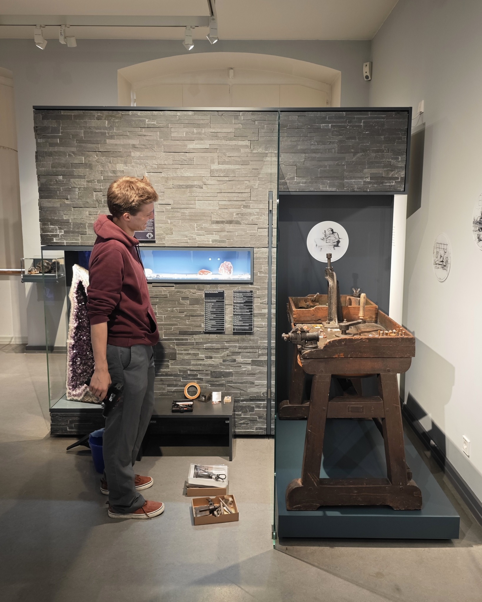 Eine Person in einem Hoodie und grauer Hose betrachtet in einem Museum eine alte Holzwerkbank mit Werkzeugen. Links daneben steht eine Vitrine mit einem großen Kristall, auf dem Boden liegen weitere Werkzeugkästen. Im Hintergrund ist eine graue Steinwand mit Infotafeln zu sehen. (KI-Alternativtext)