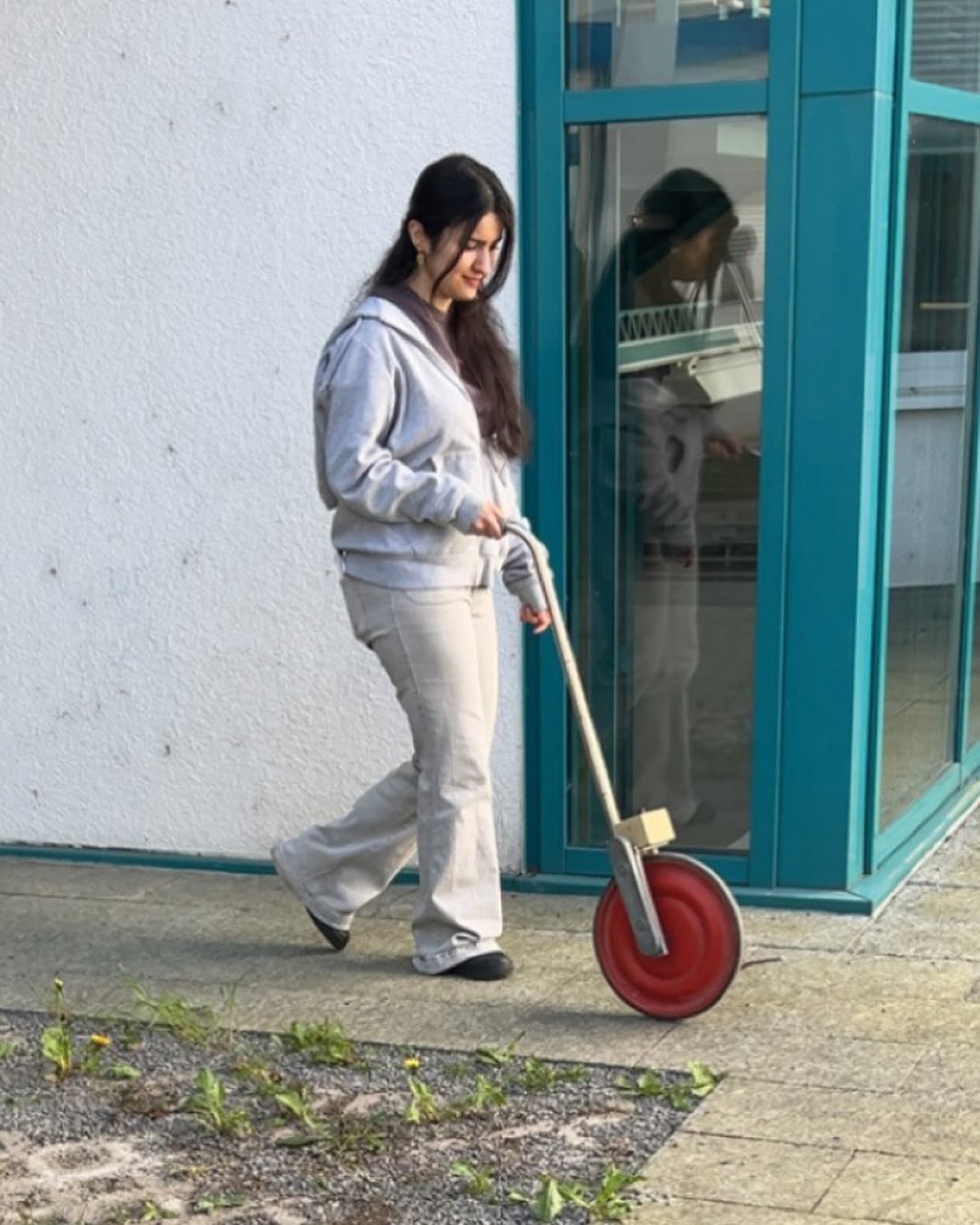 Dieser Alternativtext wurde KI-generiert: Eine Person schiebt ein zweirädriges Fahrzeug vor einem Gebäudeeingang. Das Bild ist farbig und zeigt eine Rückansicht.