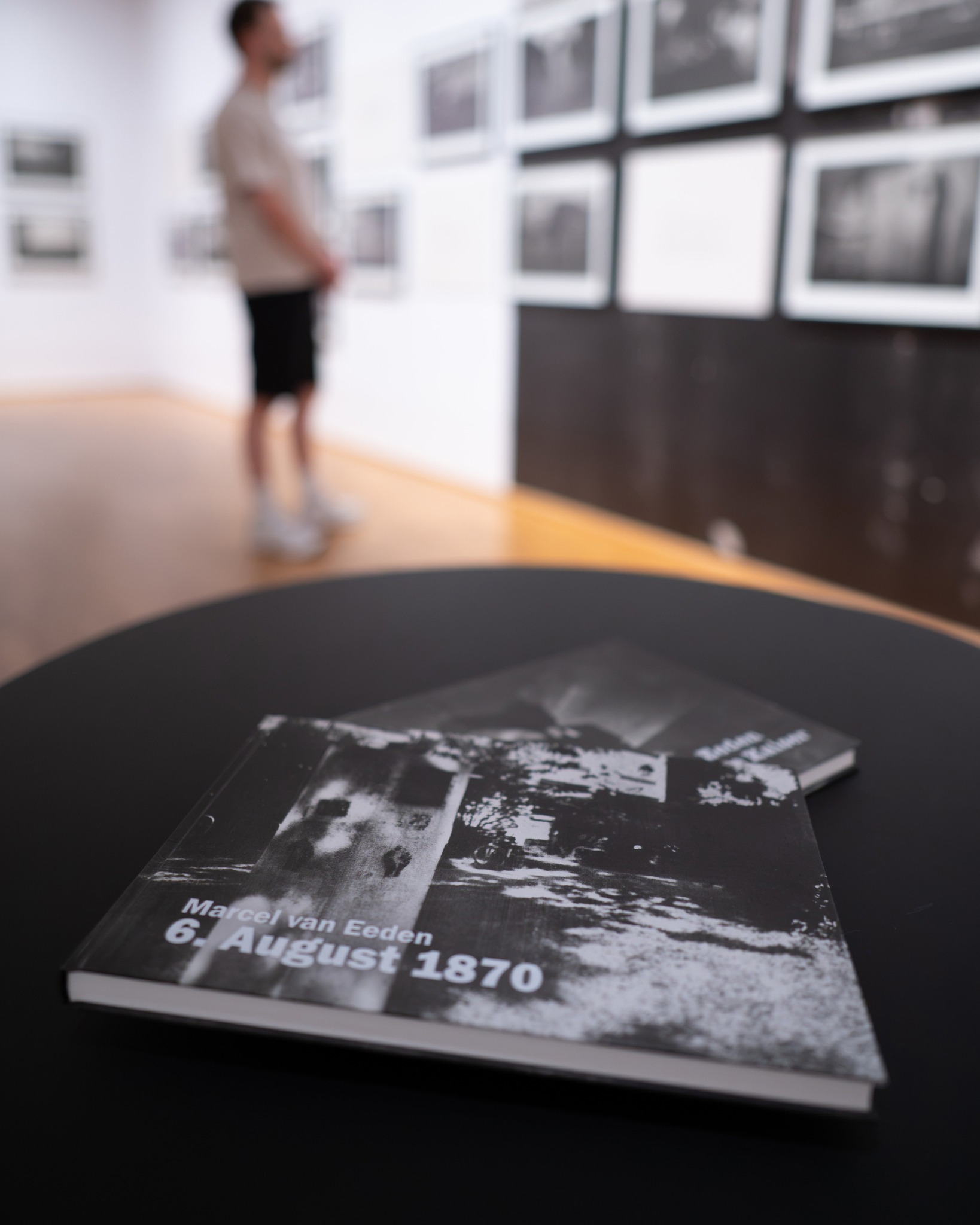Auf einem schwarzen Tisch liegen zwei Bücher, wobei das vordere Buch den Titel „Marcel van Eeden 6. August 1870“ trägt. Im unscharfen Hintergrund steht eine Person in einer Galerie und betrachtet eine Wand voller gerahmter Schwarz-Weiß-Fotografien. (KI-Alternativtext)