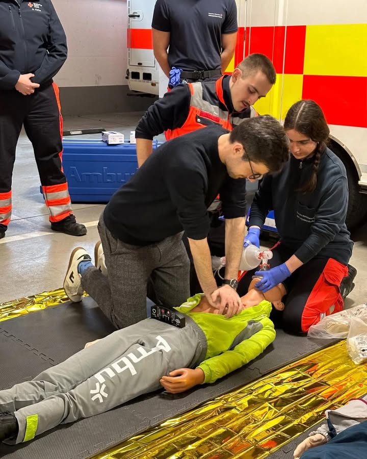 Dieser Alternativtext wurde KI-generiert: Mehrere Personen in Einsatzkleidung beugen sich über eine liegende Person auf einer Rettungsmatte. Im Hintergrund ist ein Rettungswagen zu sehen.