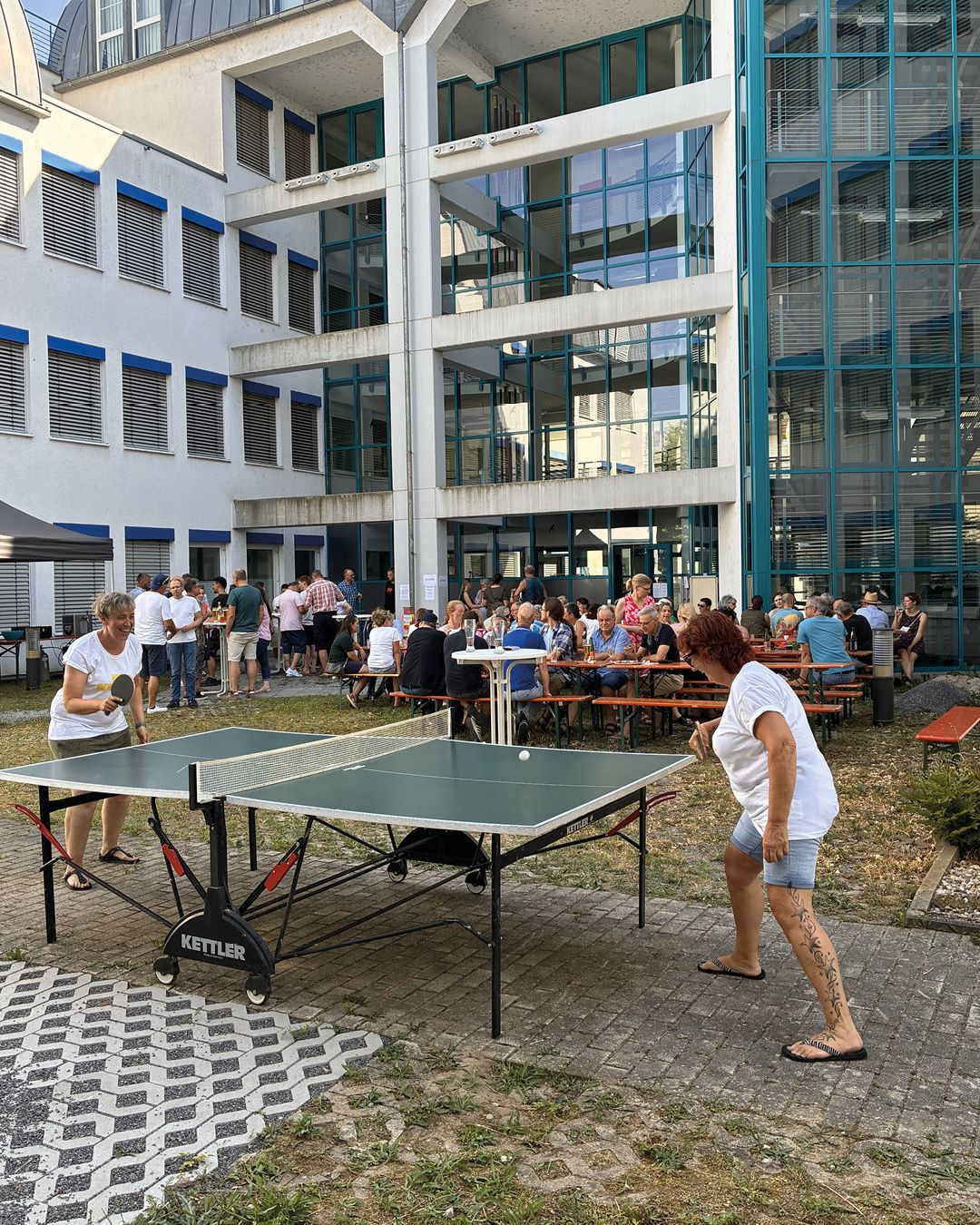 Zwei Personen spielen auf einem gepflasterten Platz Tischtennis an einem grünen Tisch mit der Aufschrift KETTLER. Im Hintergrund sitzen und stehen zahlreiche Personen an Holztischen auf einer Wiese vor einem modernen Gebäude mit großer Glasfassade. (KI-Alternativtext)