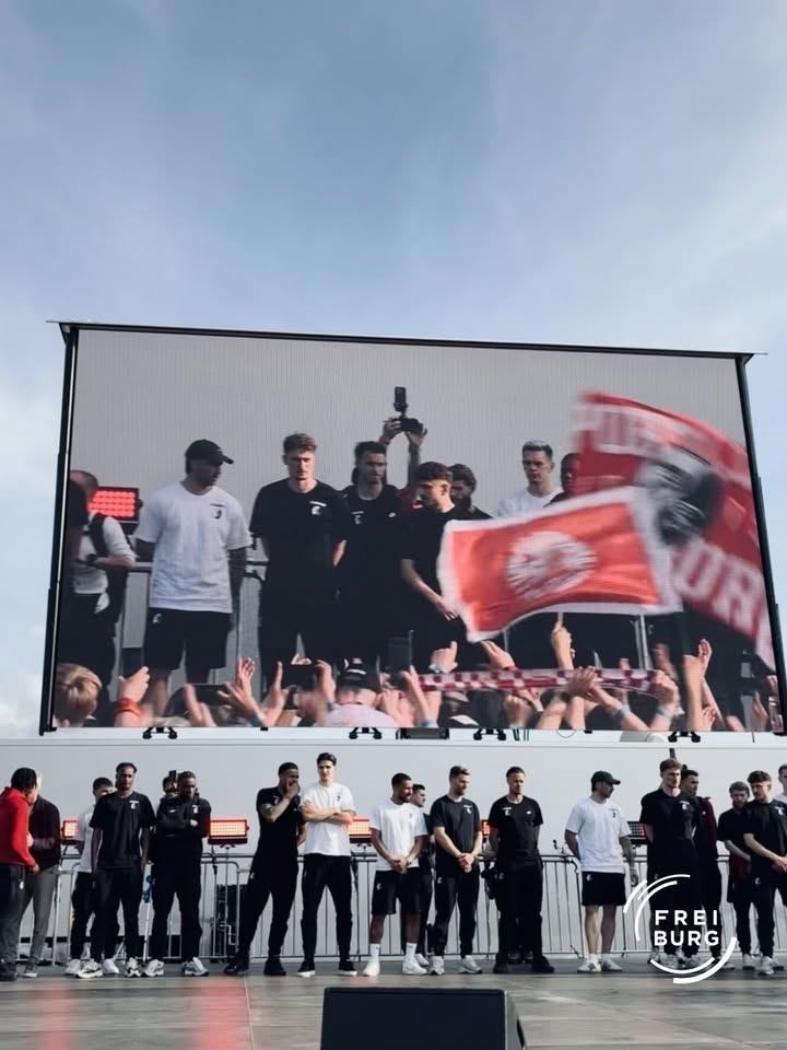 Mehrere Personen stehen auf einer Bühne vor einem großen Bildschirm, der eine Gruppe mit roten Fahnen und jubelndem Publikum zeigt. Einige tragen dunkle oder weiße Sportkleidung, eine Person hält ein Smartphone hoch. Im Vordergrund ist das Logo „Freiburg“ sichtbar. (KI-Alternativtext)