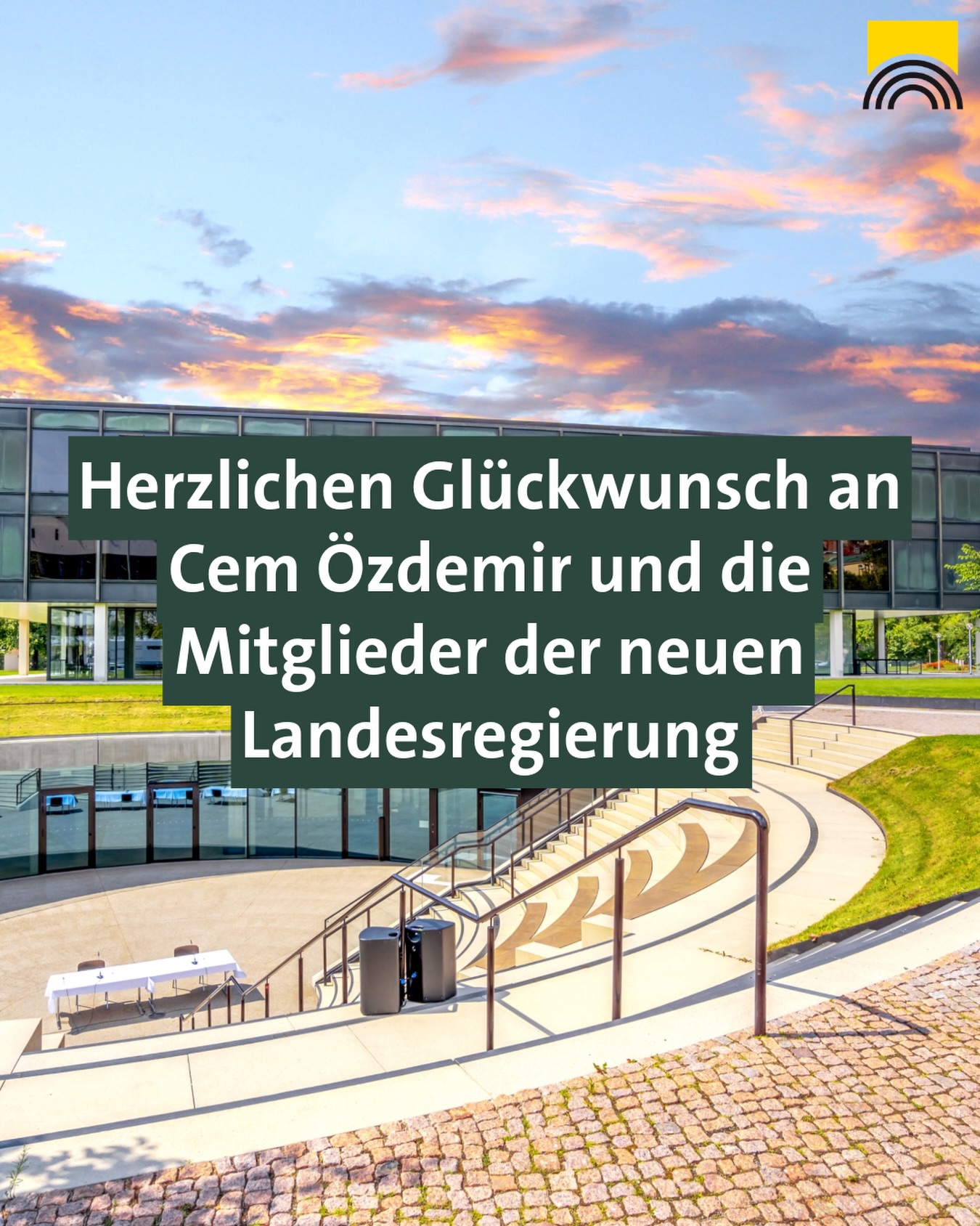 Herzlichen Glückwunsch an Cem Özdemir und die Mitglieder der neuen Landesregierung. Im Hintergrund modernes Gebäude mit Treppen und Tisch unter einem farbenfrohen Himmel, rechts oben Logo mit gelbem Quadrat und Regenbogenbogen. (KI-Alternativtext)